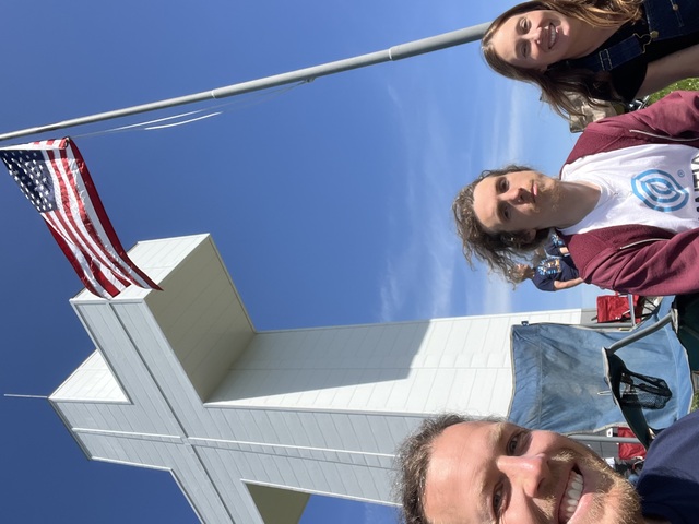 Devon, Charles, and Erica, in front of a USA flag and the Bald Knob Cross of Peace